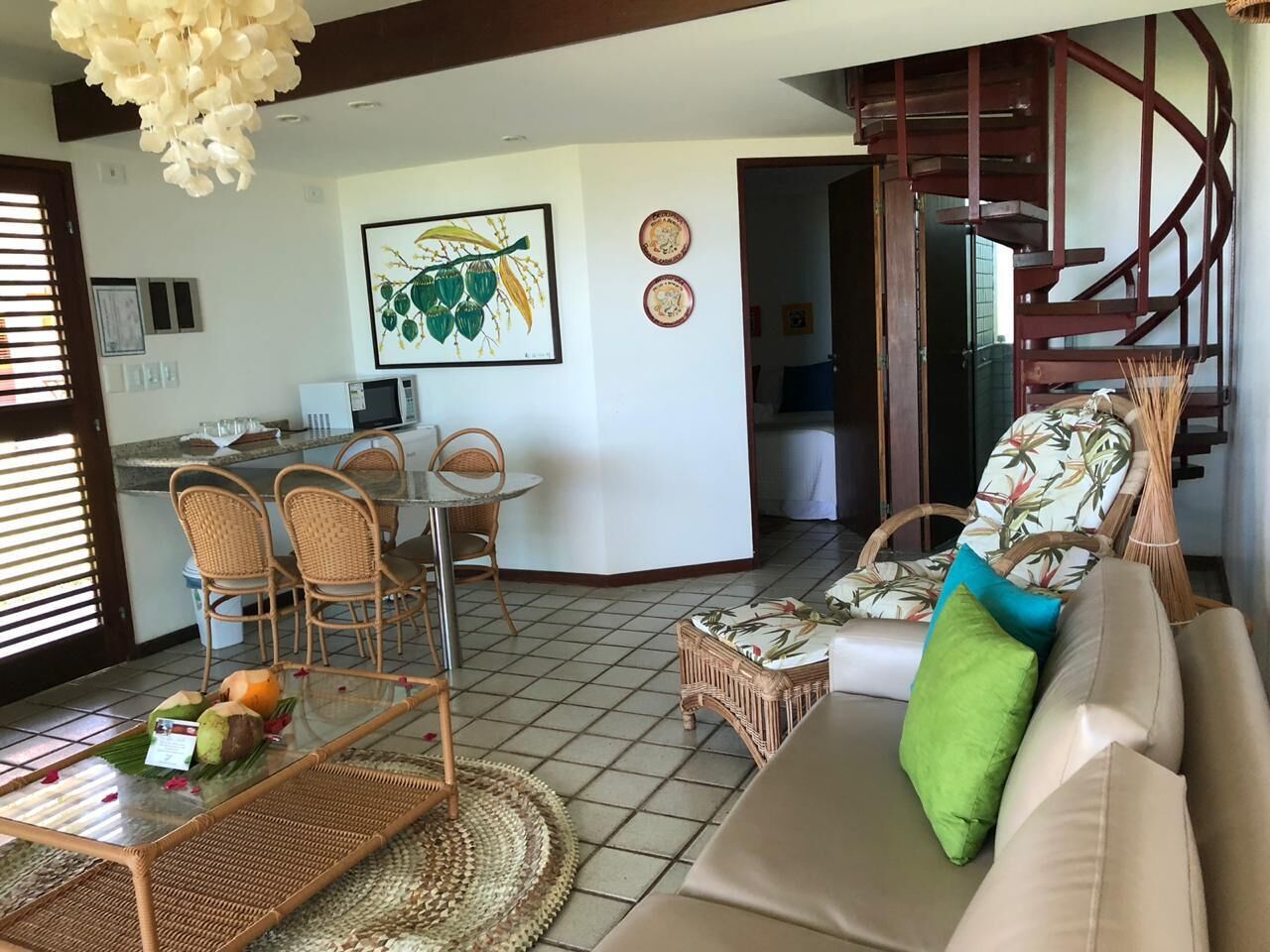 Un salon avec des meubles en osier, un canapé agrémenté d’un coussin vert, un coin repas avec des chaises, une cuisine en arrière-plan et un escalier en colimaçon.