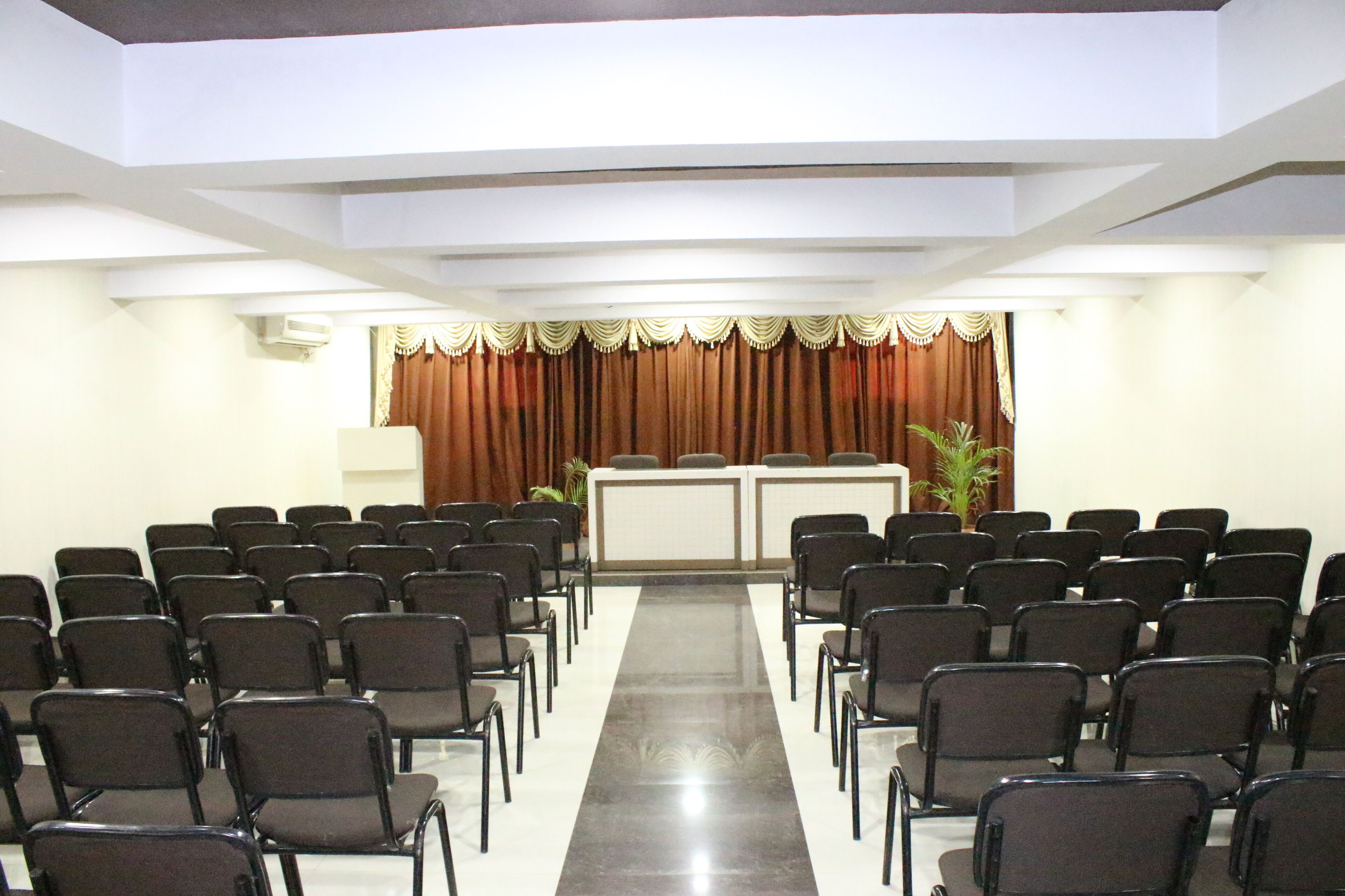 A conference room with rows of chairs facing a podium, white walls, and a decorative curtain.