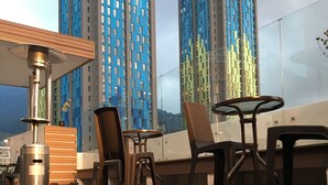 Una terraza en la azotea con mobiliario exterior, un basurero y un calentador. Al fondo, edificios altos con fachadas azules y verdes.