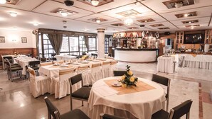 Un restaurante con mesas redondas, manteles blancos y sillas. Cuenta con una zona de bar con mostrador y estanterías llenas de botellas.