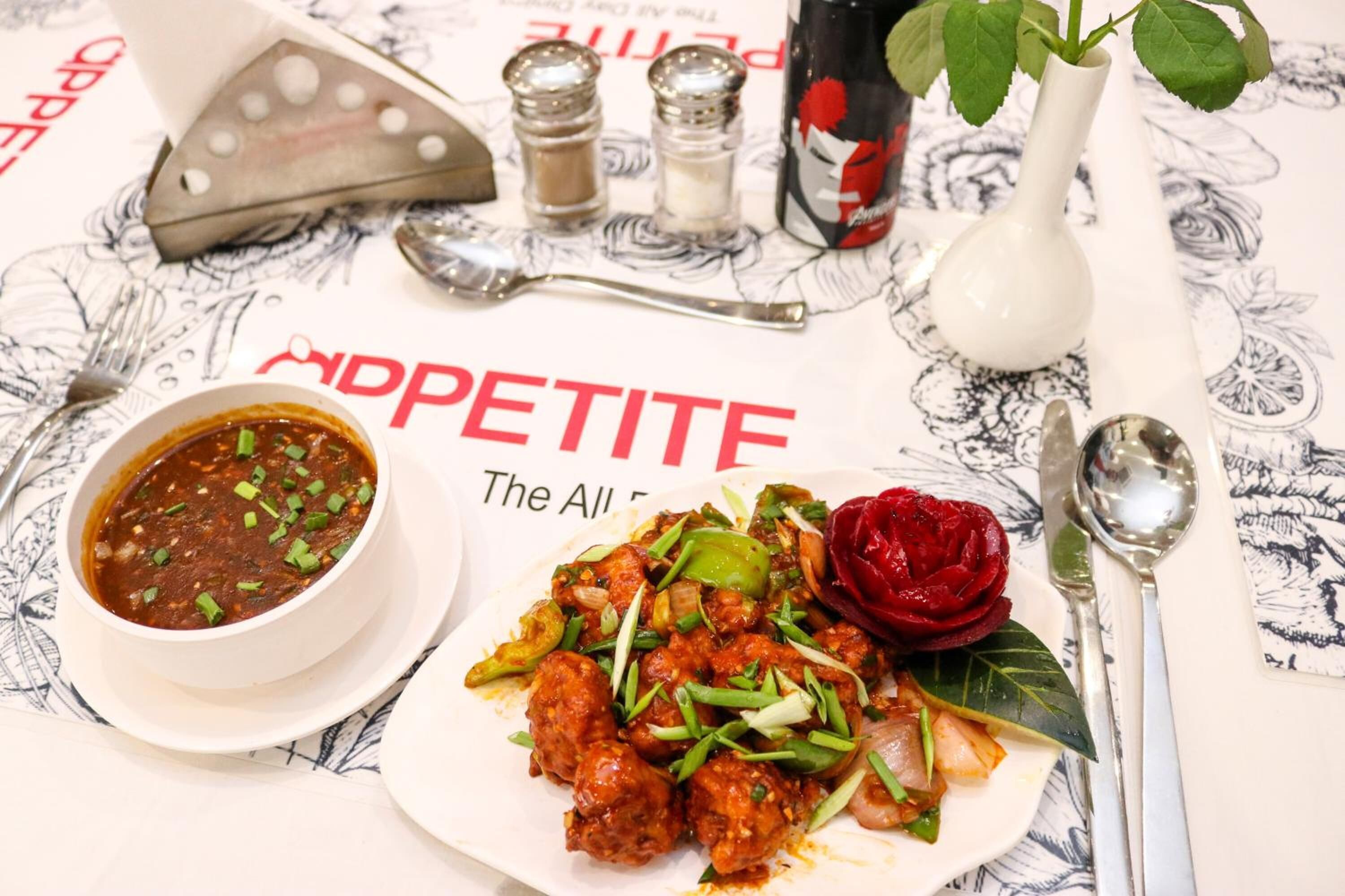 Una mesa con un plato de sopa en un bol, vegetales salteados en un plato, y una flor roja. Hay saleros, pimenteros, una botella y una cuchara sobre la mesa.