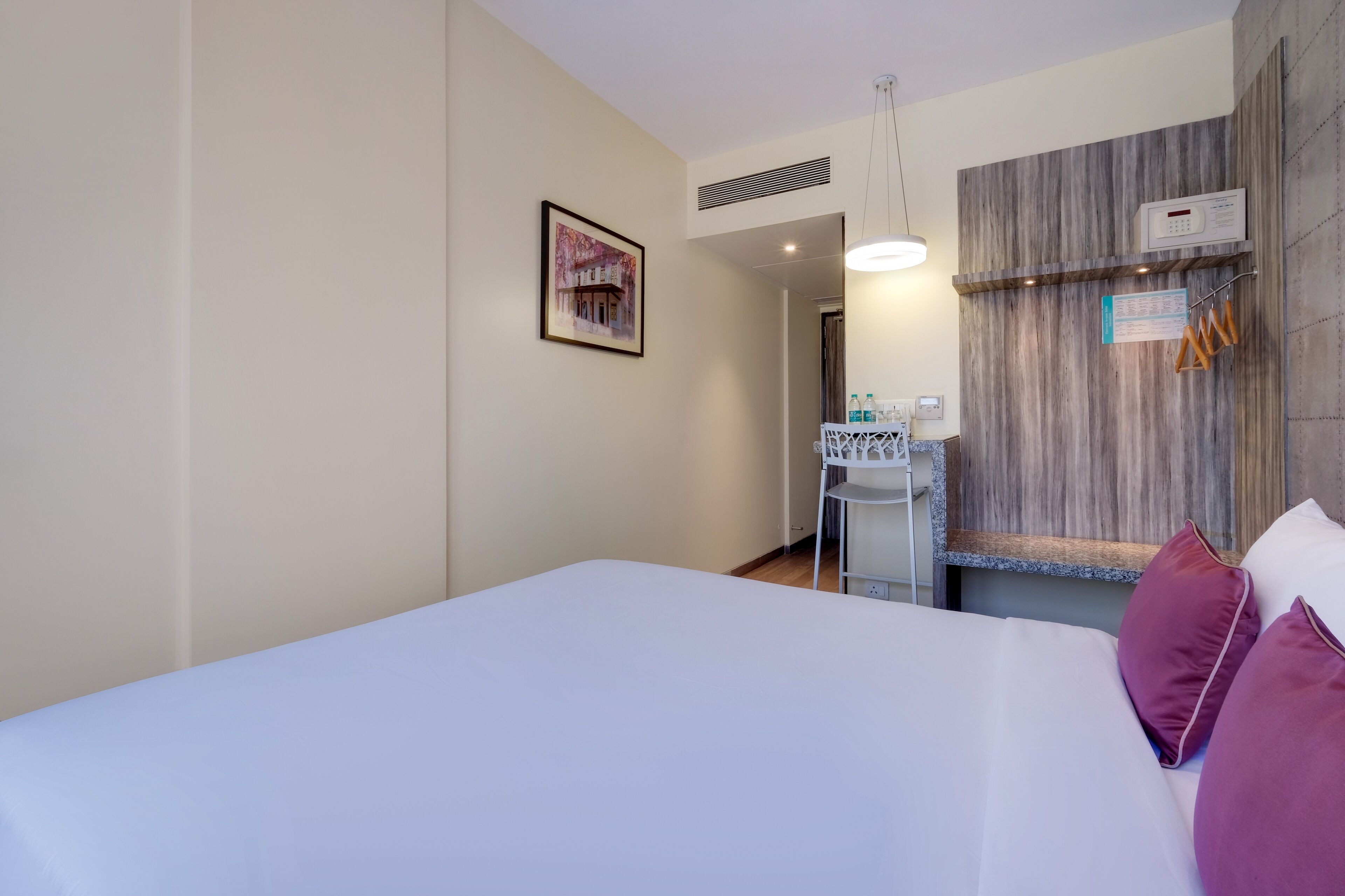 Una camera d'albergo con un letto, una piccola scrivania con una sedia, uno scaffale a parete con articoli da toilette e un quadro appeso.