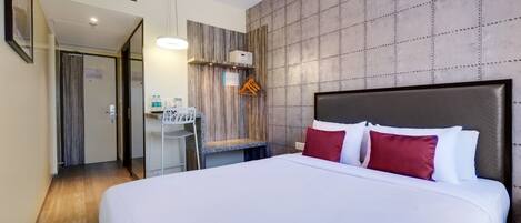 A hotel room with a large bed, a desk with a chair, a hanging light fixture, and a wardrobe.