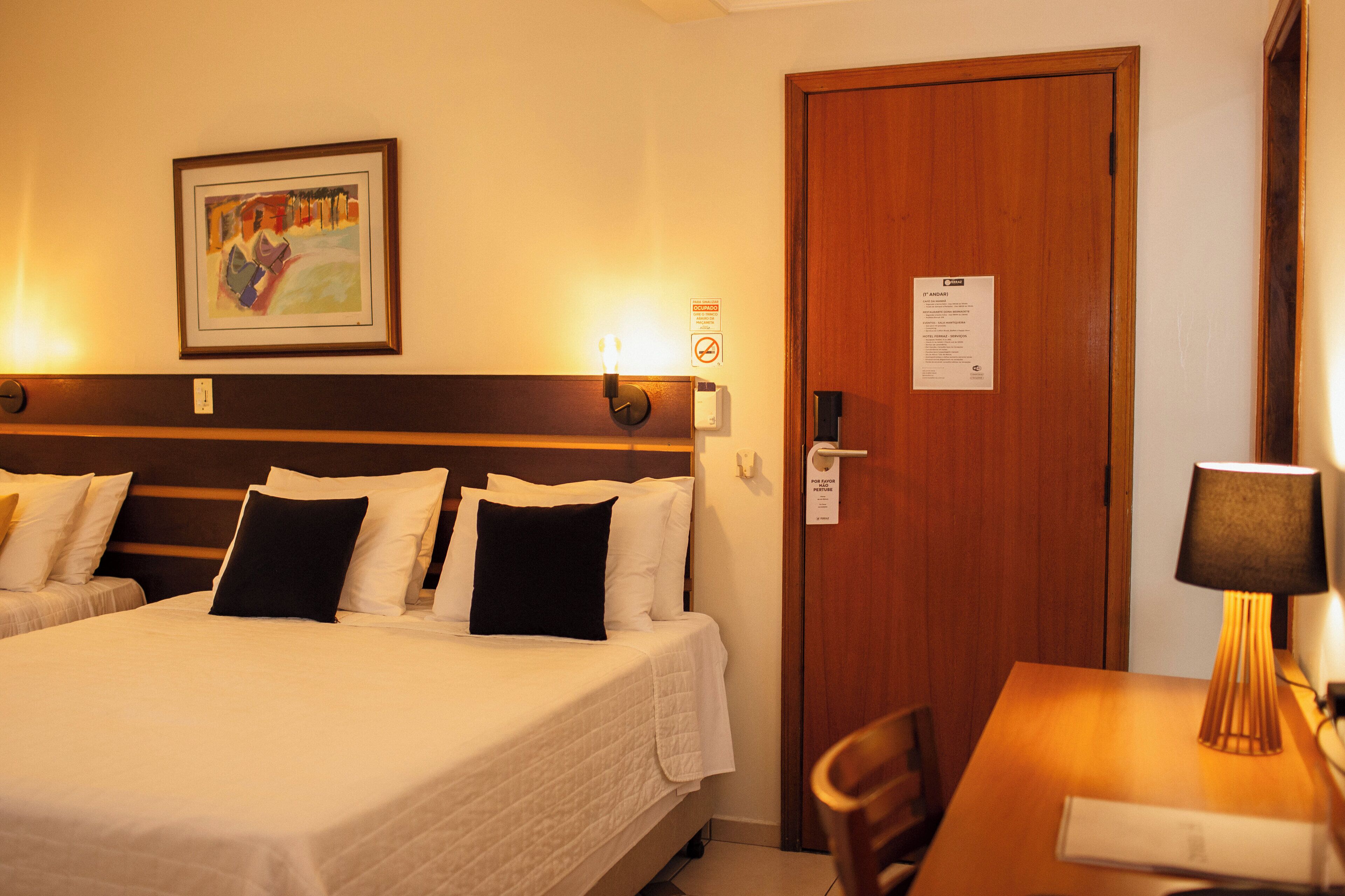 Une chambre d’hôtel avec un grand lit, un bureau et une chaise. Il y a un tableau au mur et une porte avec une pancarte.