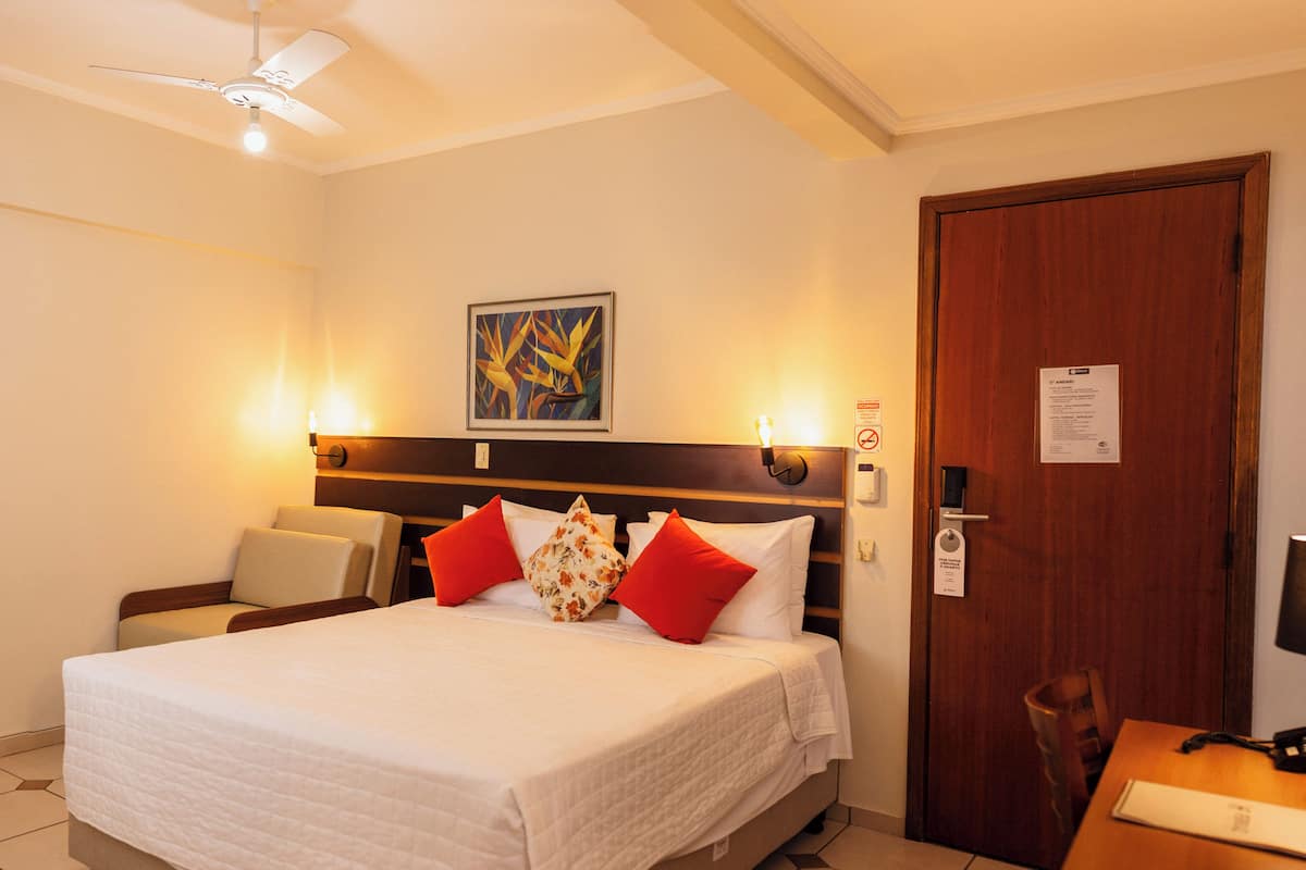 A hotel room with a bed, a desk with a lamp, a chair, a door, and a ceiling fan.