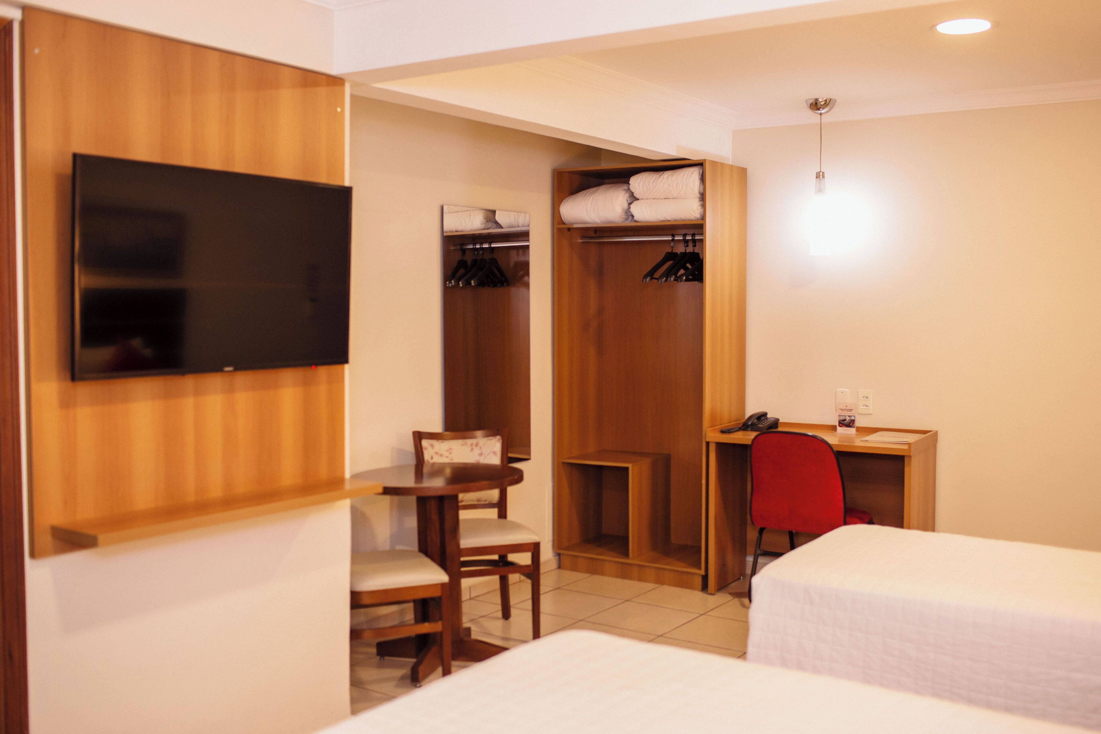 A hotel room with two beds, a desk with a chair, a TV, a closet with hanging clothes, and a small table with a chair.