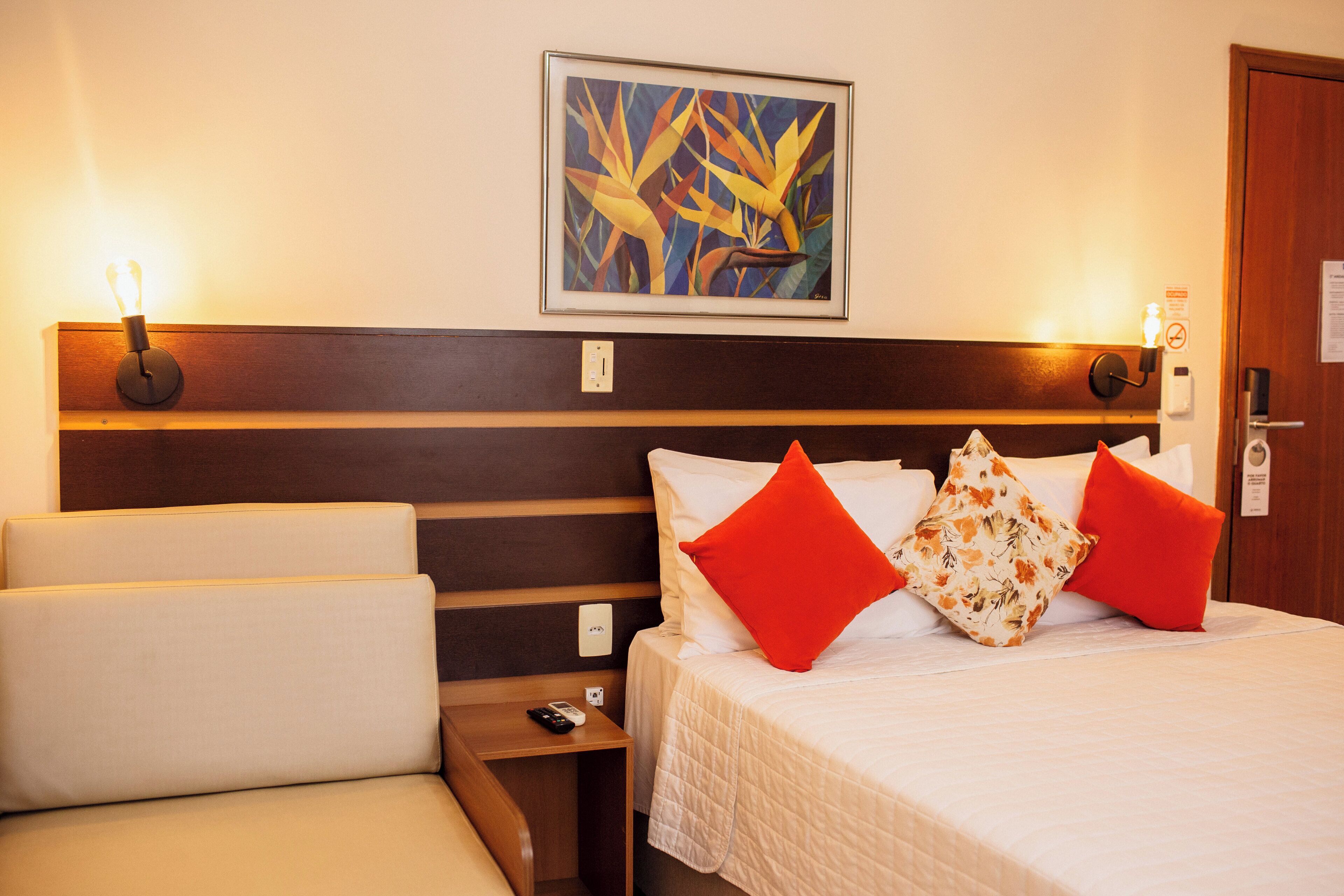 Quarto de hotel com cabeceira de madeira, cama com roupa de cama branca e almofadas vermelhas e florais, mesa de cabeceira com controle remoto e quadro pendurado na parede.