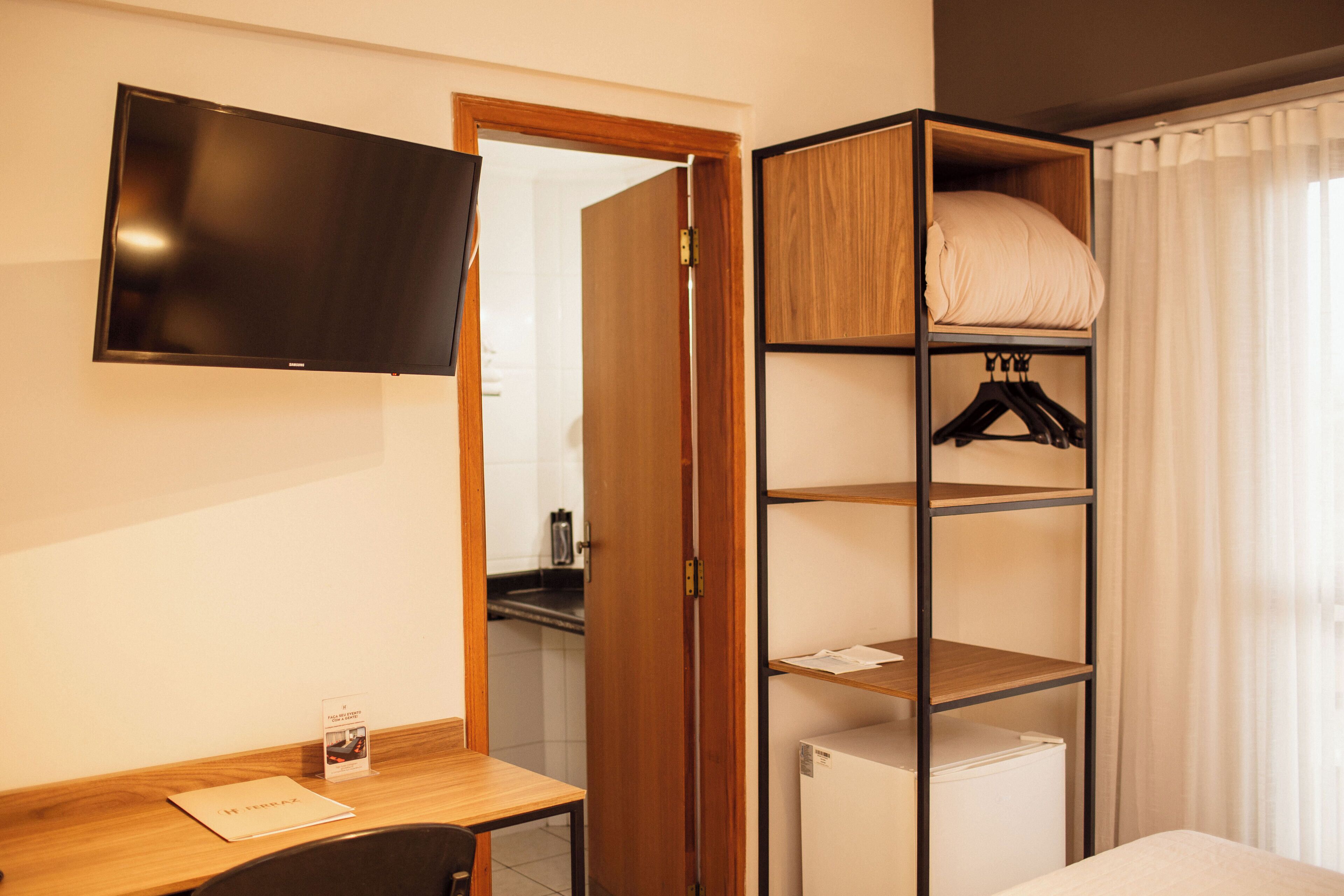 Habitación de hotel con un escritorio de madera, un televisor de pantalla plana montado en la pared, un armario con estantes y una cama con edredón blanco.
