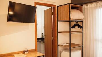 Habitación de hotel con un escritorio de madera, un televisor de pantalla plana montado en la pared, un armario con estantes y una cama con edredón blanco.