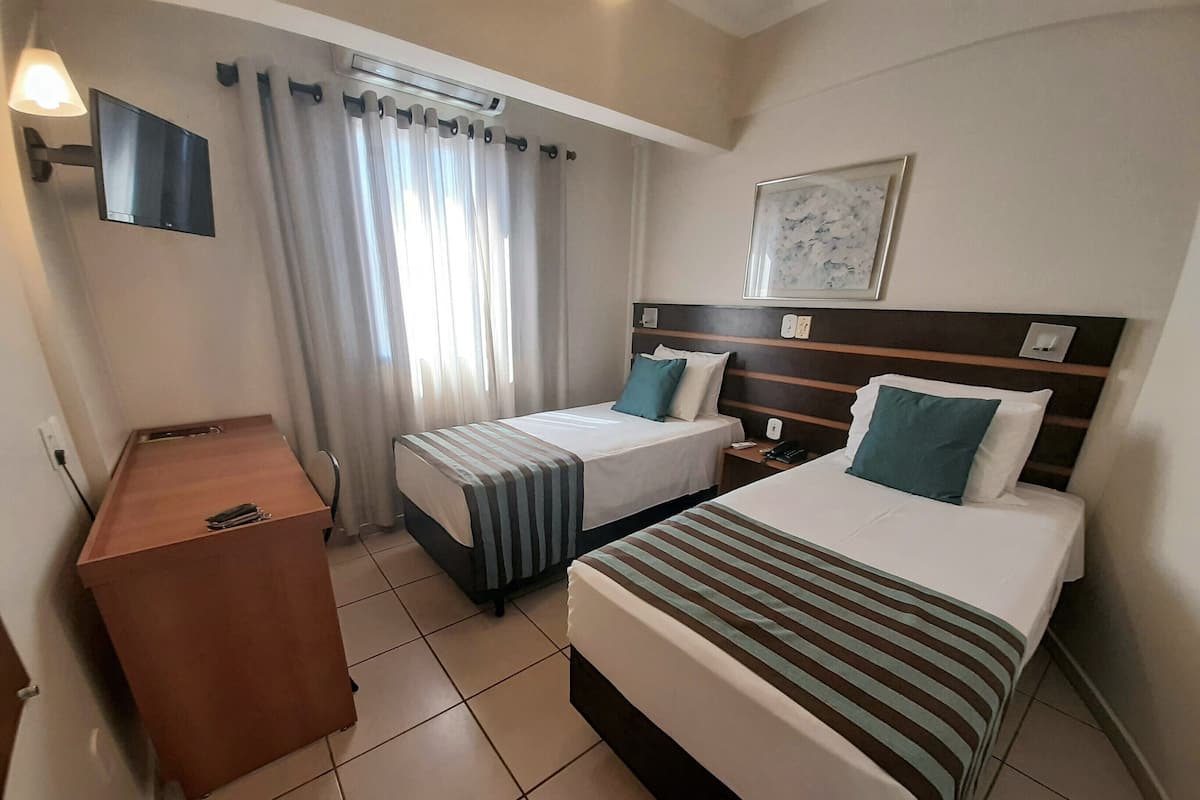 A hotel room with two beds, a desk, a chair, a flat-screen TV, and a window with curtains.