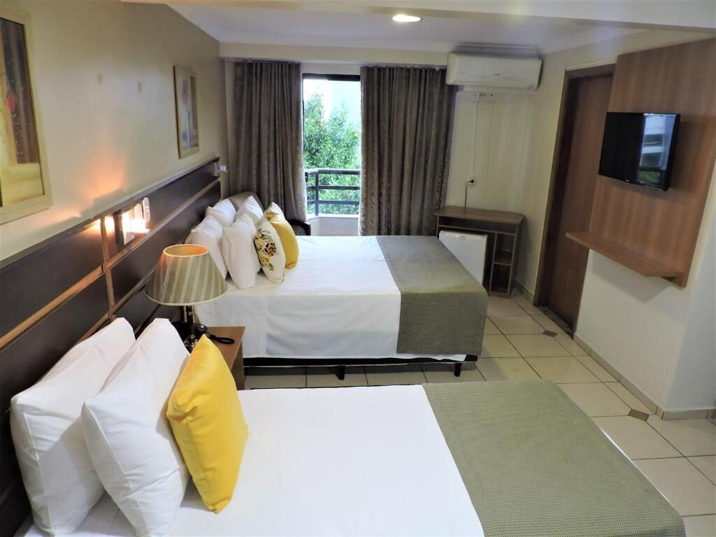 A hotel room with two beds, a desk, a TV, and a balcony with curtains.