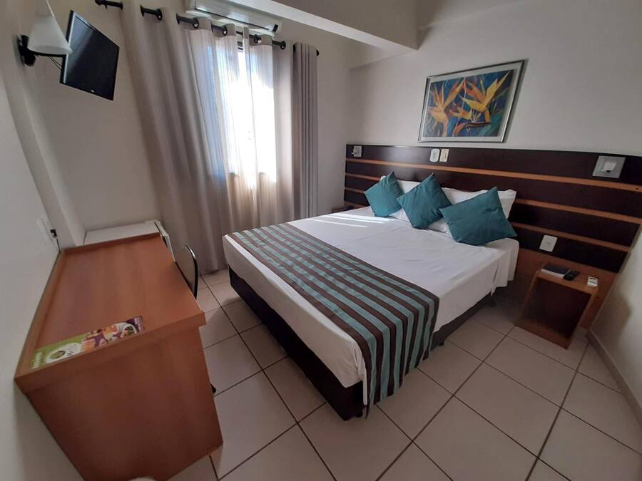 A hotel room with a bed, a desk, a chair, a TV, and a painting on the wall.