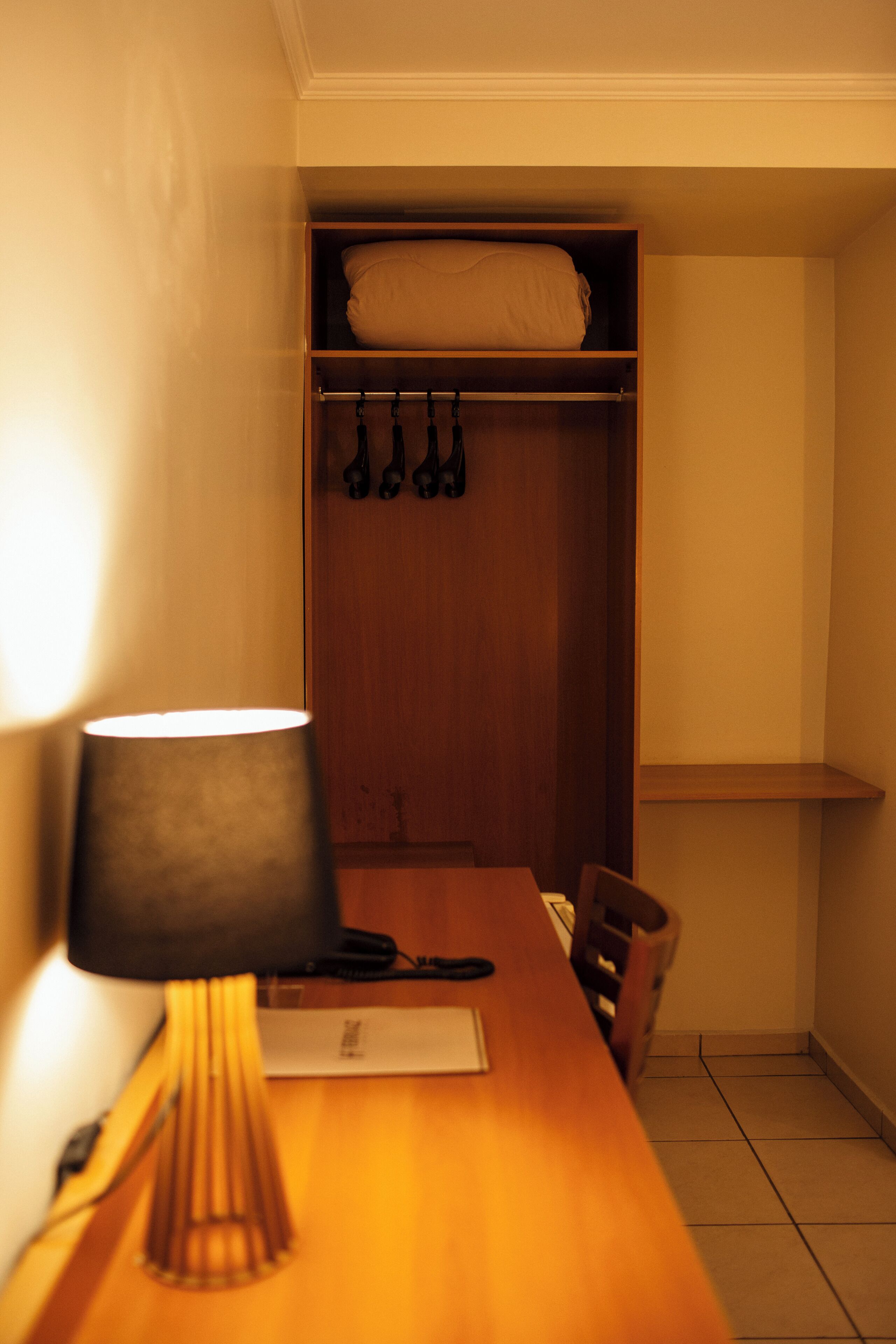 A hotel room with a wooden desk, a lamp, a chair, a closet with hanging items, and a tiled floor.