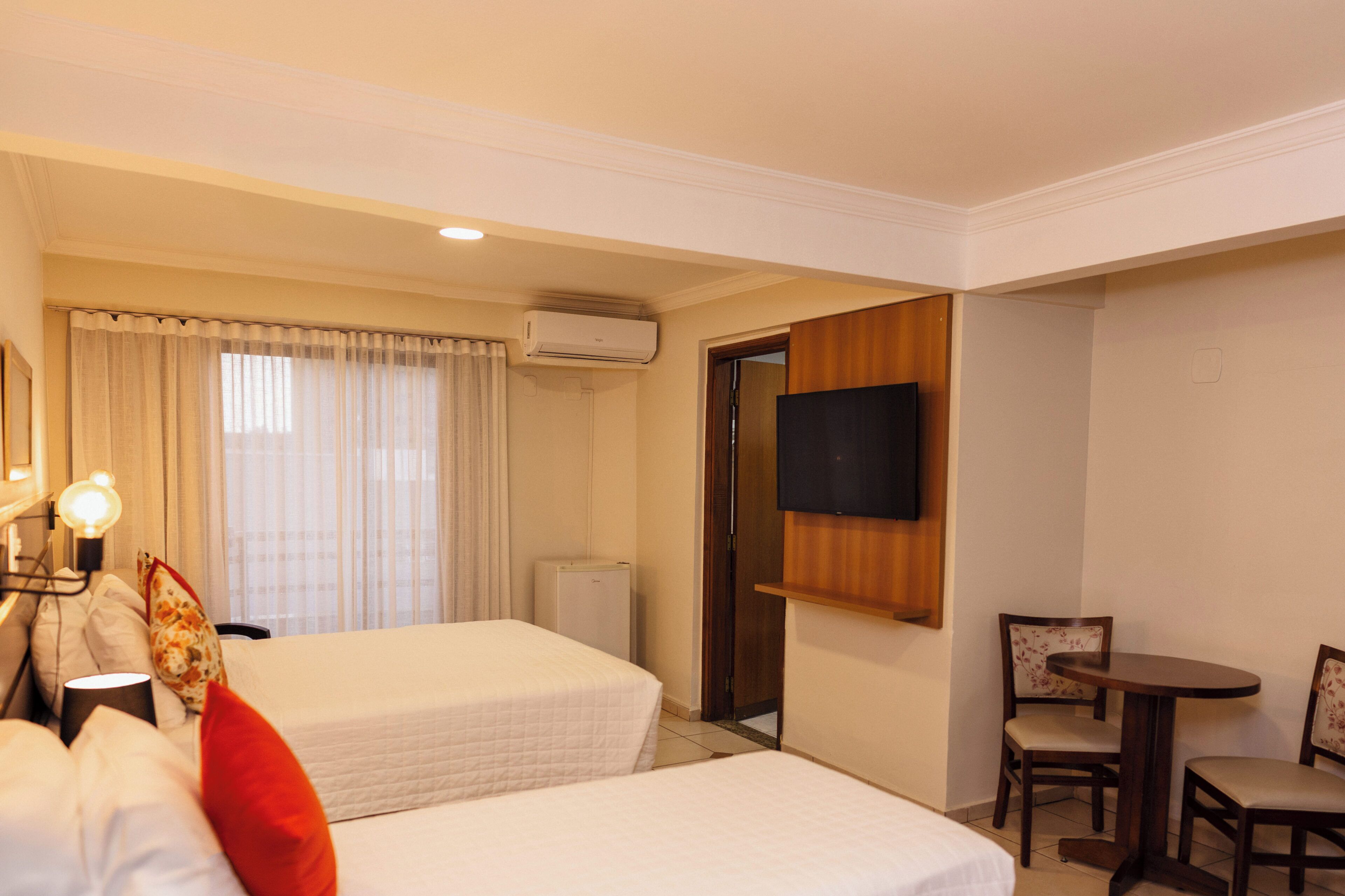 A hotel room with two beds, a TV mounted on the wall, a small table with chairs, and an air conditioning unit.