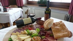 A charcuterie board with various cheeses, meats, and bread, accompanied by a bottle of wine and a glass of red wine, set on a table in a room with a window and curtains. - Hotel Col Di Lana (Canazei)