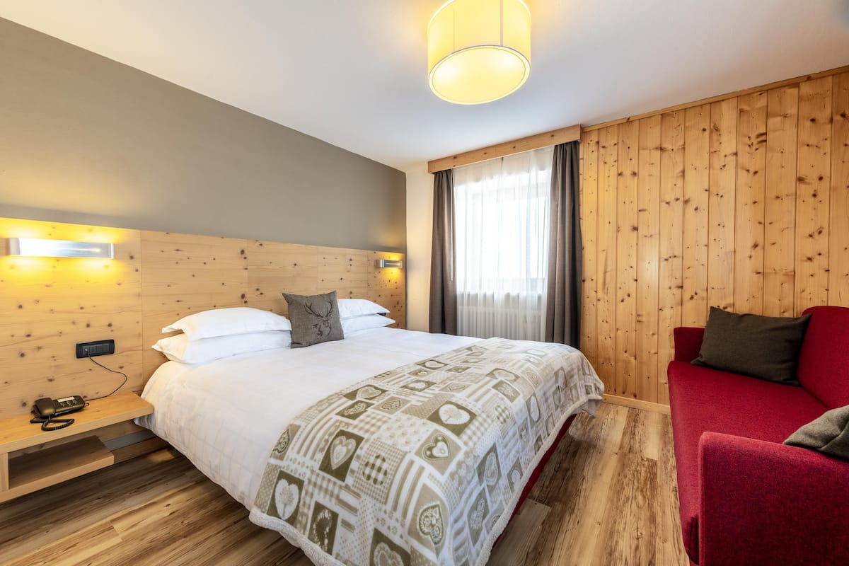 Hotellihuone, jossa on puinen sängynpääty, kuviollinen peitto sängyllä, punainen sohva ja yöpöytä, jossa on puhelin.