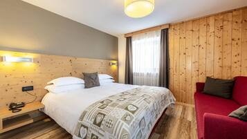 Una habitación de hotel con cabecera de madera, cama con manta estampada, sofá rojo y mesita de noche con teléfono.