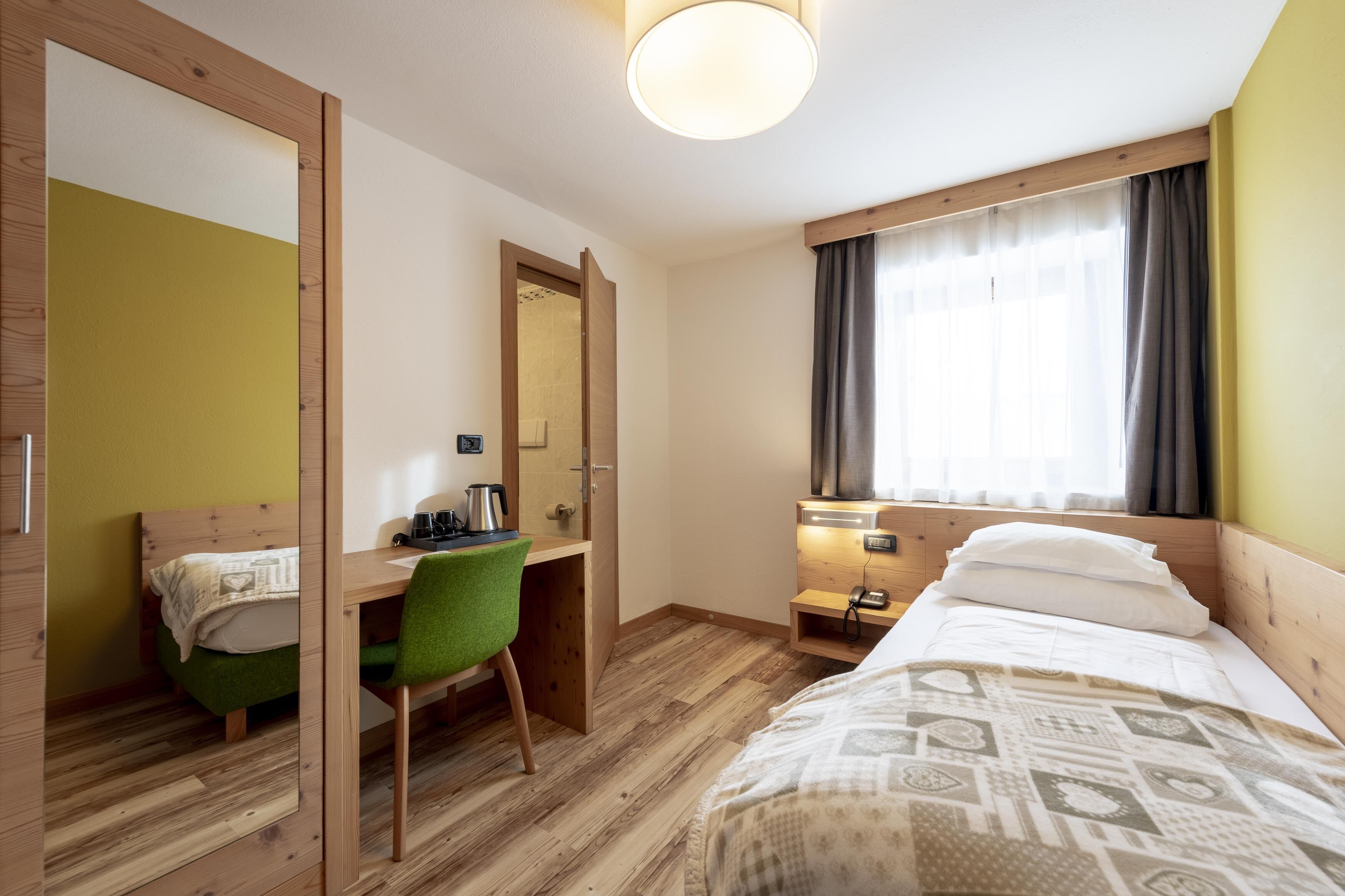 A hotel room with a bed, a desk with a chair, a mirror, a bedside table with a lamp, and a window with curtains.