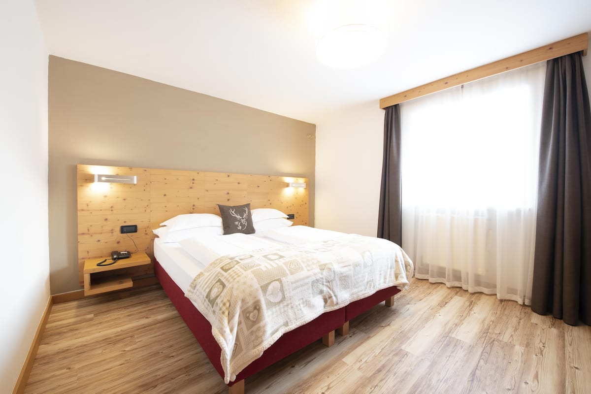 Hotellihuone, jossa on puinen sängynpääty, kuviollinen päiväpeite sängyllä, yöpöytä, jossa on puhelin, ja ikkuna, jossa on verhot.