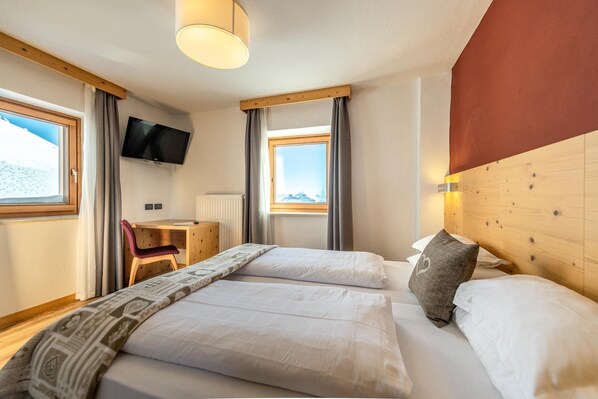 A hotel room with two beds, a desk, a chair, a TV, and a window with a view of the mountains. - Hotel Col Di Lana (Canazei)