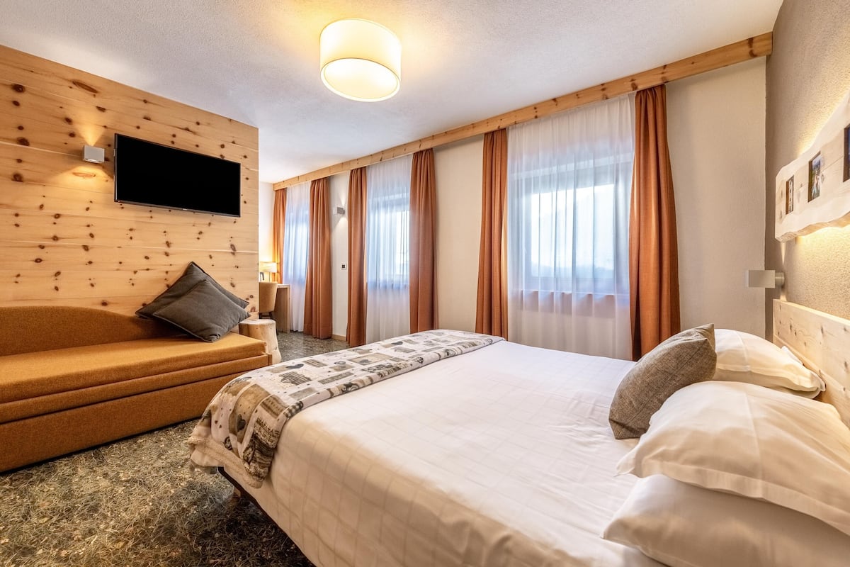 Hotellihuone, jossa on puinen seinä, taulutelevisio, valkoisilla lakanoilla varustettu sänky ja tyynyillä koristeltu sohva.