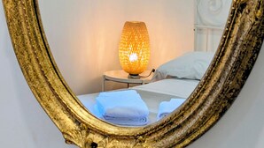 Uma cama com roupa de cama branca, um abajur e um espelho com moldura dourada refletindo o quarto.