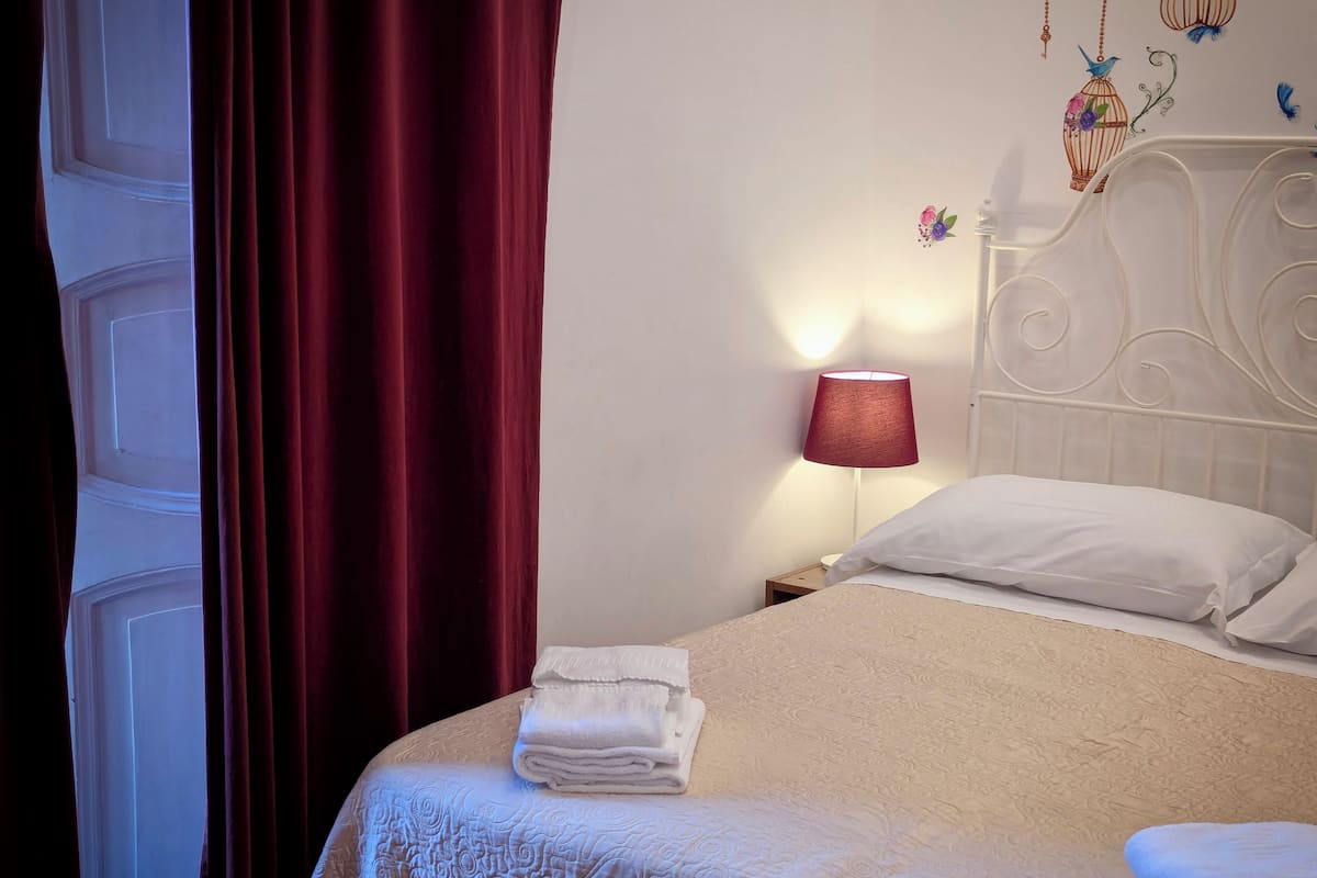 A neatly made bed with white linens, a red curtain partially drawn, and a wall with decorative bird and flower wall art.
