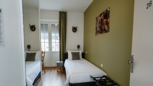 Quarto com duas camas, uma mesa pequena e uma cadeira. Há duas luminárias fixadas na parede e um quadro na parede.