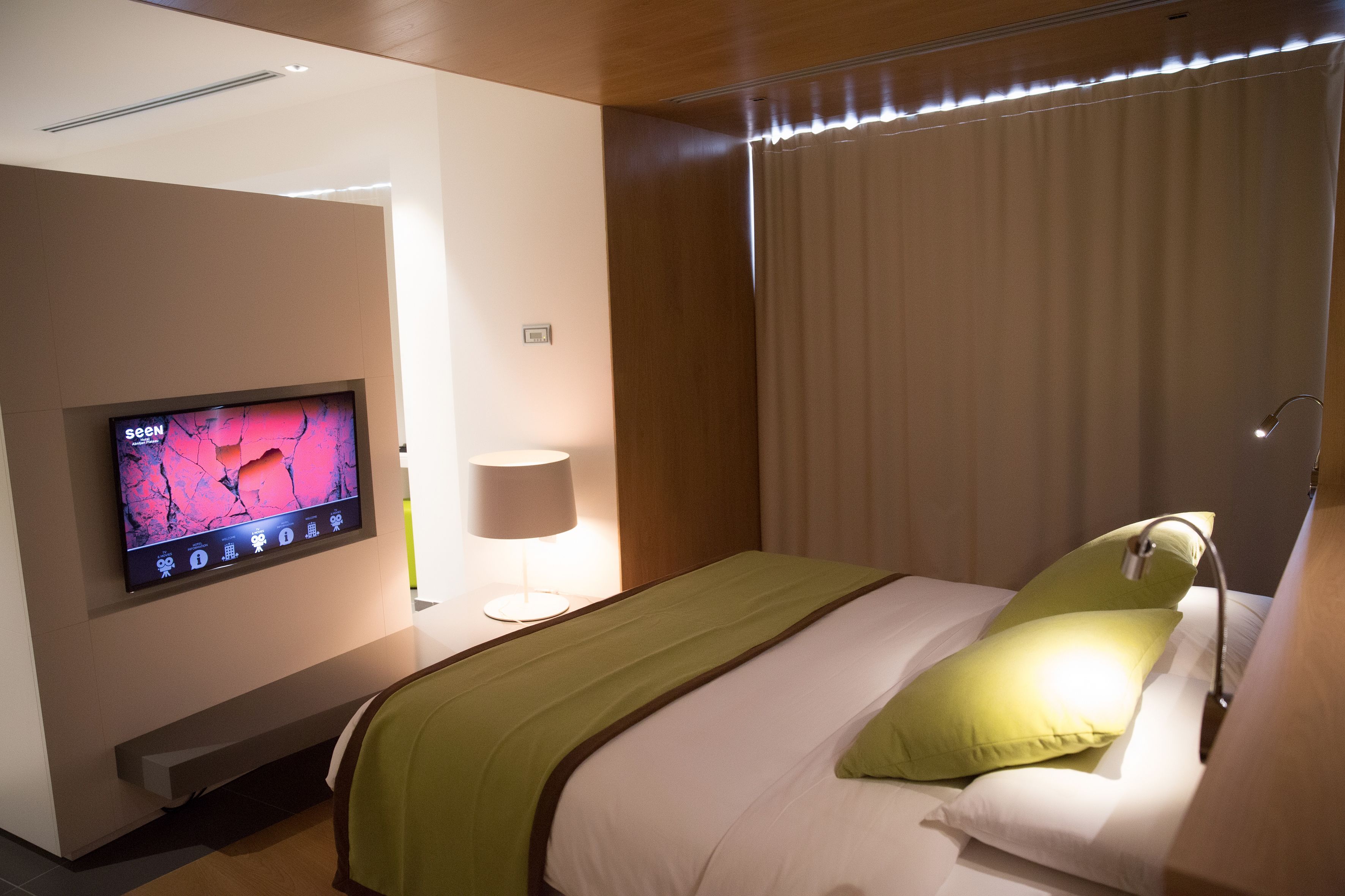 A hotel room with a bed, a TV on the wall, a lamp, and a window with curtains.