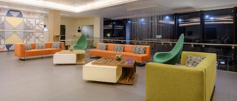 A modern lounge area with orange and green sofas, a wooden coffee table, and a glass railing overlooking a cityscape.