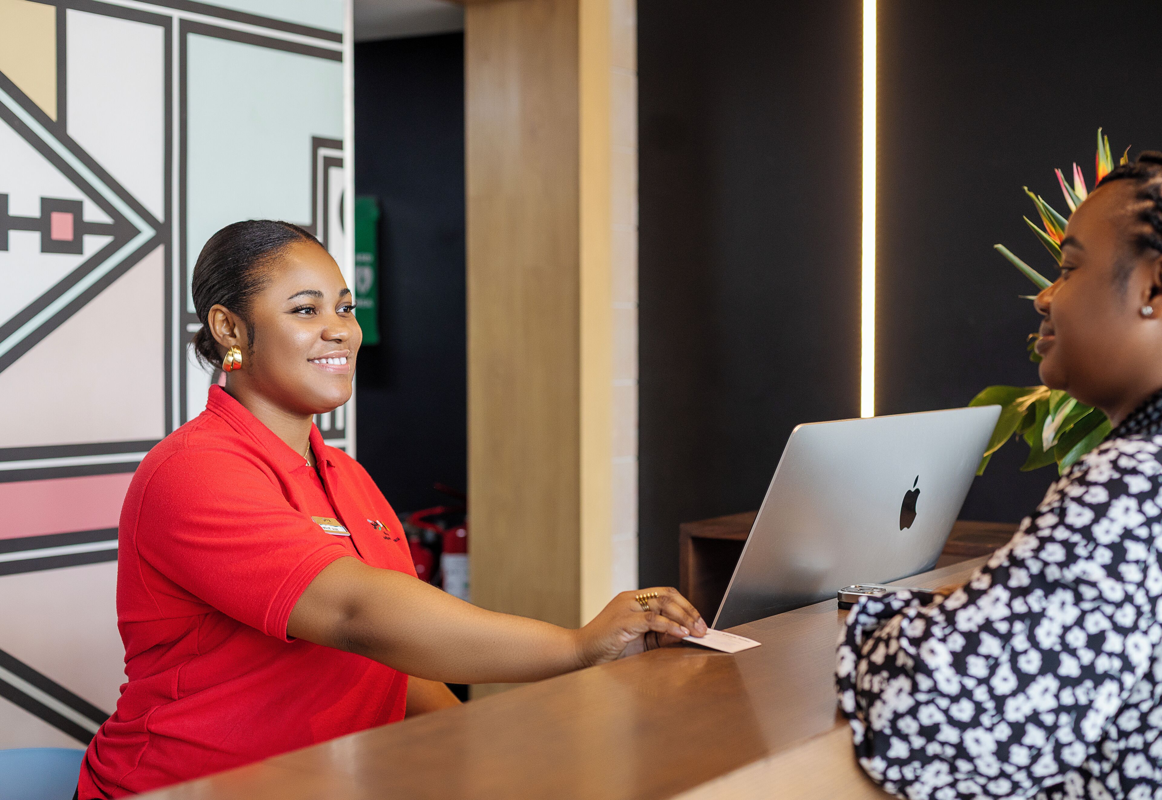 Zona de recepción con una recepcionista con uniforme rojo, un cliente con una computadora portátil y un diseño moderno de pared geométrica.