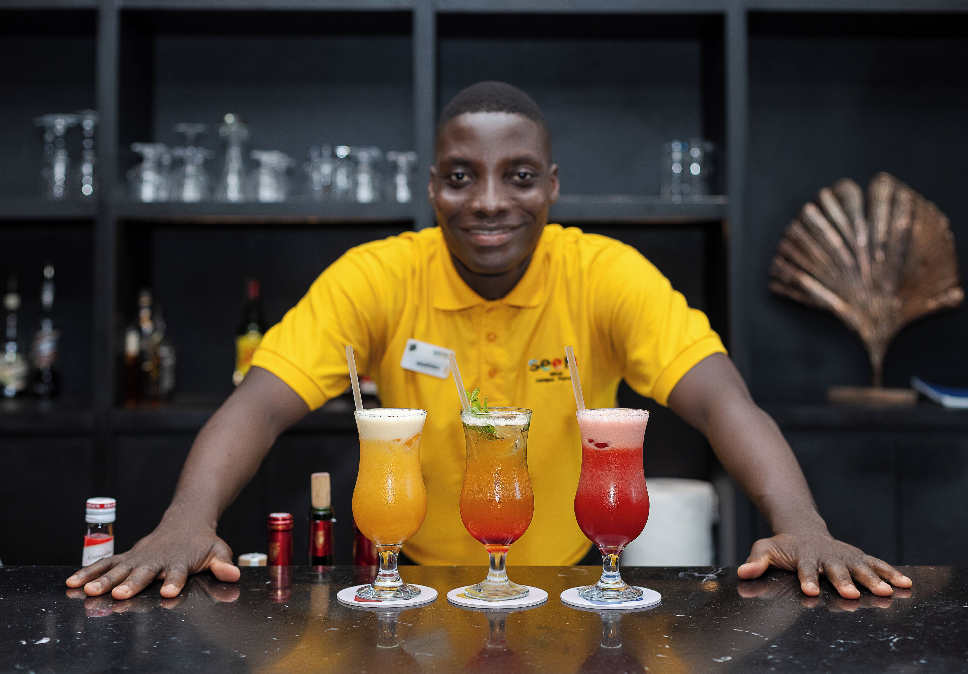 A bartender in a yellow shirt presenting three colorful cocktails on a bar counter.