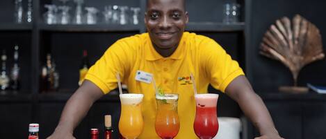 A bartender in a yellow shirt presenting three colorful cocktails on a bar counter.