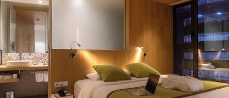A modern hotel room with a large bed, a laptop, a bedside phone, and a view of the city through a large window.