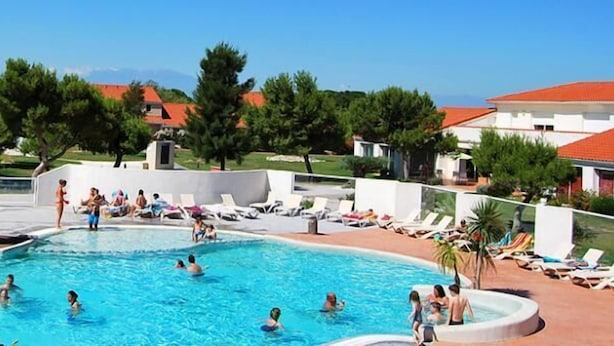 Un espace piscine où des personnes nagent et se détendent, entouré de bâtiments aux toits recouverts de tuiles rouges et de verdure.