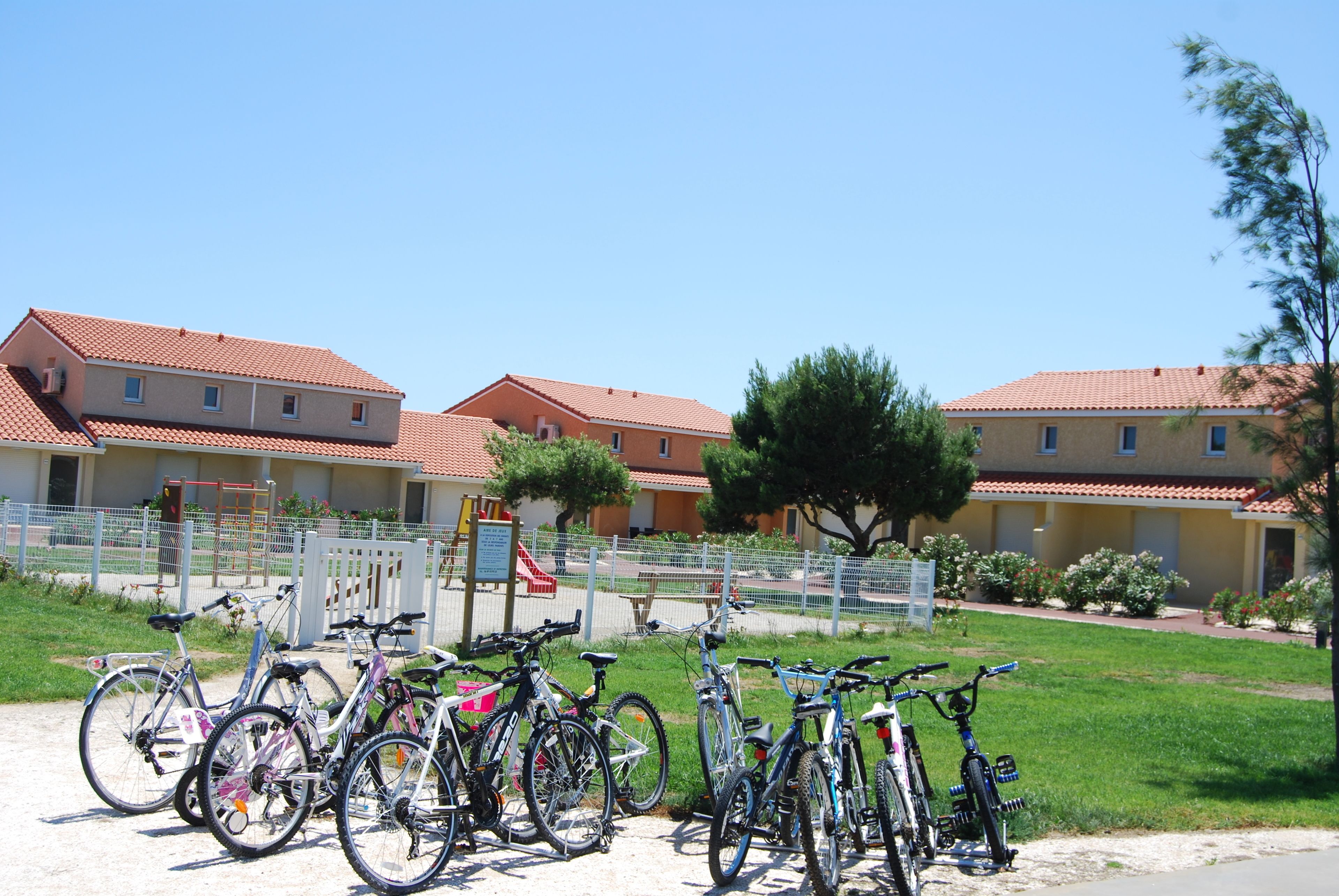 A row of bicycles parked in front of a building with a red-tiled roof and a fenced playground area.