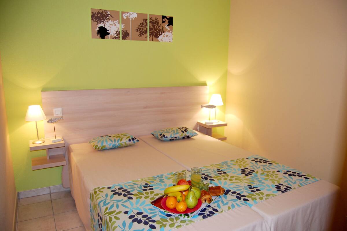Hotelkamer met een bed, twee bedlampen, een schaal fruit op het bed en een muur met drie ingelijste foto's.