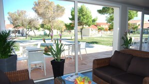 Un espace extérieur moderne aménagé avec des meubles en osier, une table en verre sur laquelle sont disposés des fruits, et une vue sur une cour intérieure avec un espace de restauration en plein air et des arbres.