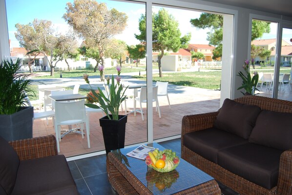 A modern outdoor seating area with wicker furniture, a glass table with fruit, and a view of a courtyard with outdoor dining and trees.