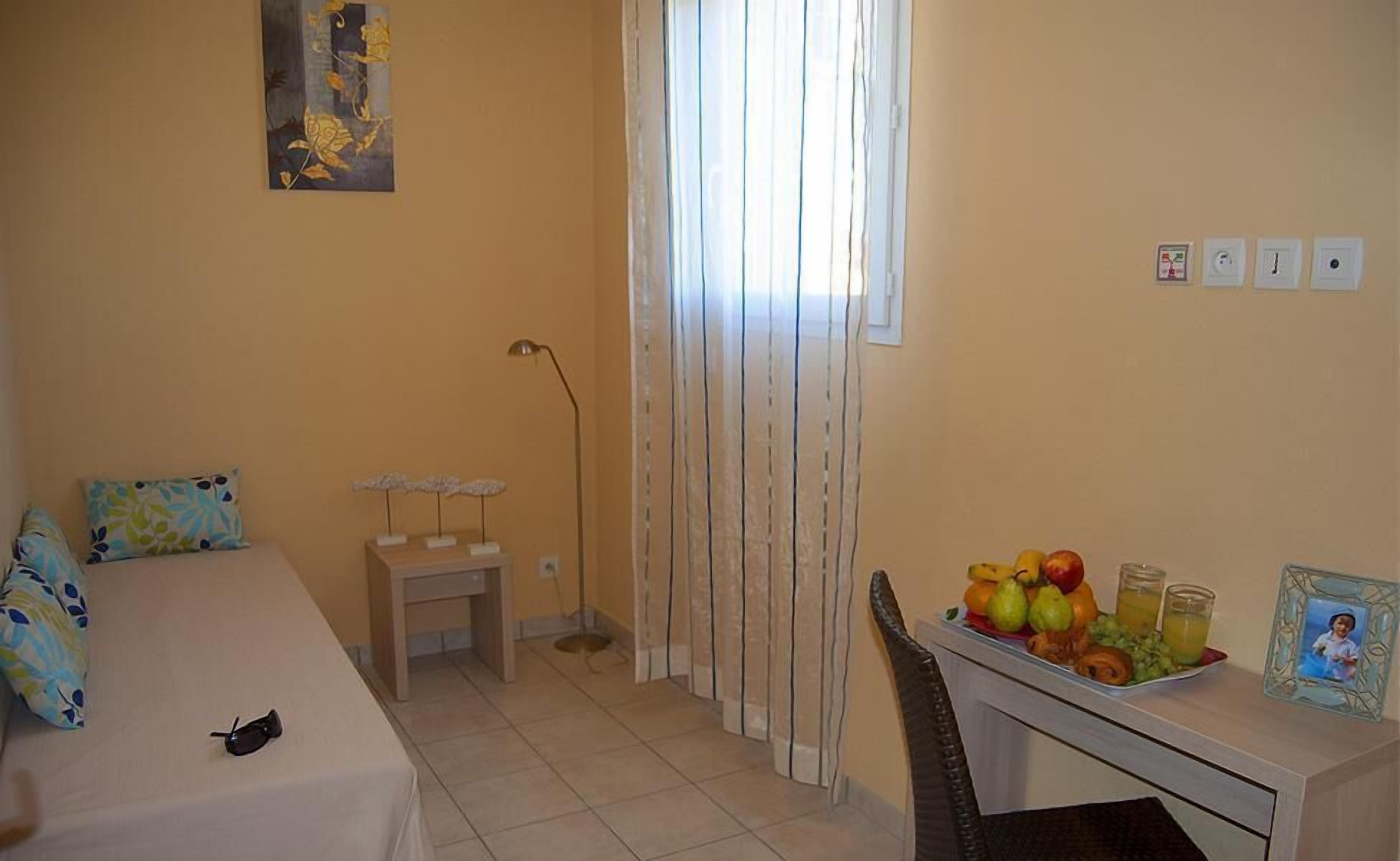 Une petite chambre à coucher, bien rangée, avec un lit, une table de chevet et un bureau sur lequel se trouvent un panier de fruits et un cadre photo.