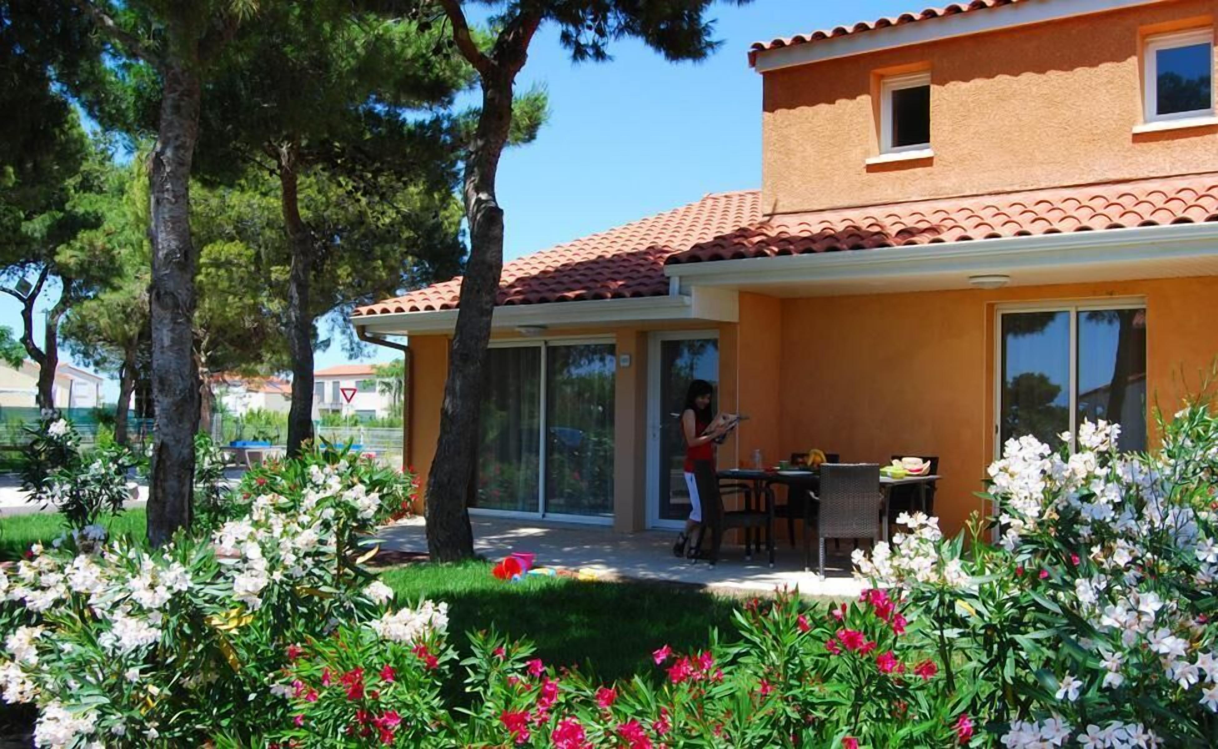 A two-story house with a tiled roof, a patio with outdoor furniture, and a garden with various flowers.