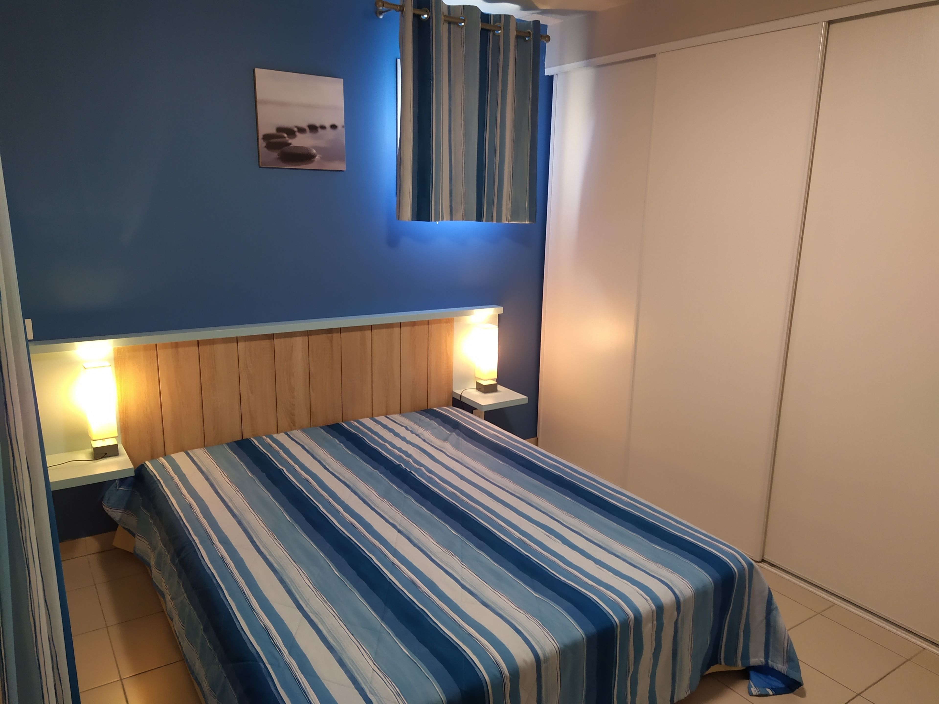 Un dormitorio con cama, dos mesitas de noche, una mesilla, una lámpara de pared, una cortina y un armario.