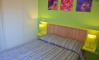 Una cama con colcha a rayas y dos lámparas de noche, cabecero de madera y tres cuadros de flores en la pared.