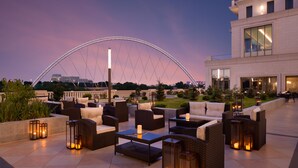 An outdoor seating area with wicker furniture and candles, overlooking a cityscape and a large bridge. - The St. Regis Astana (Astana)