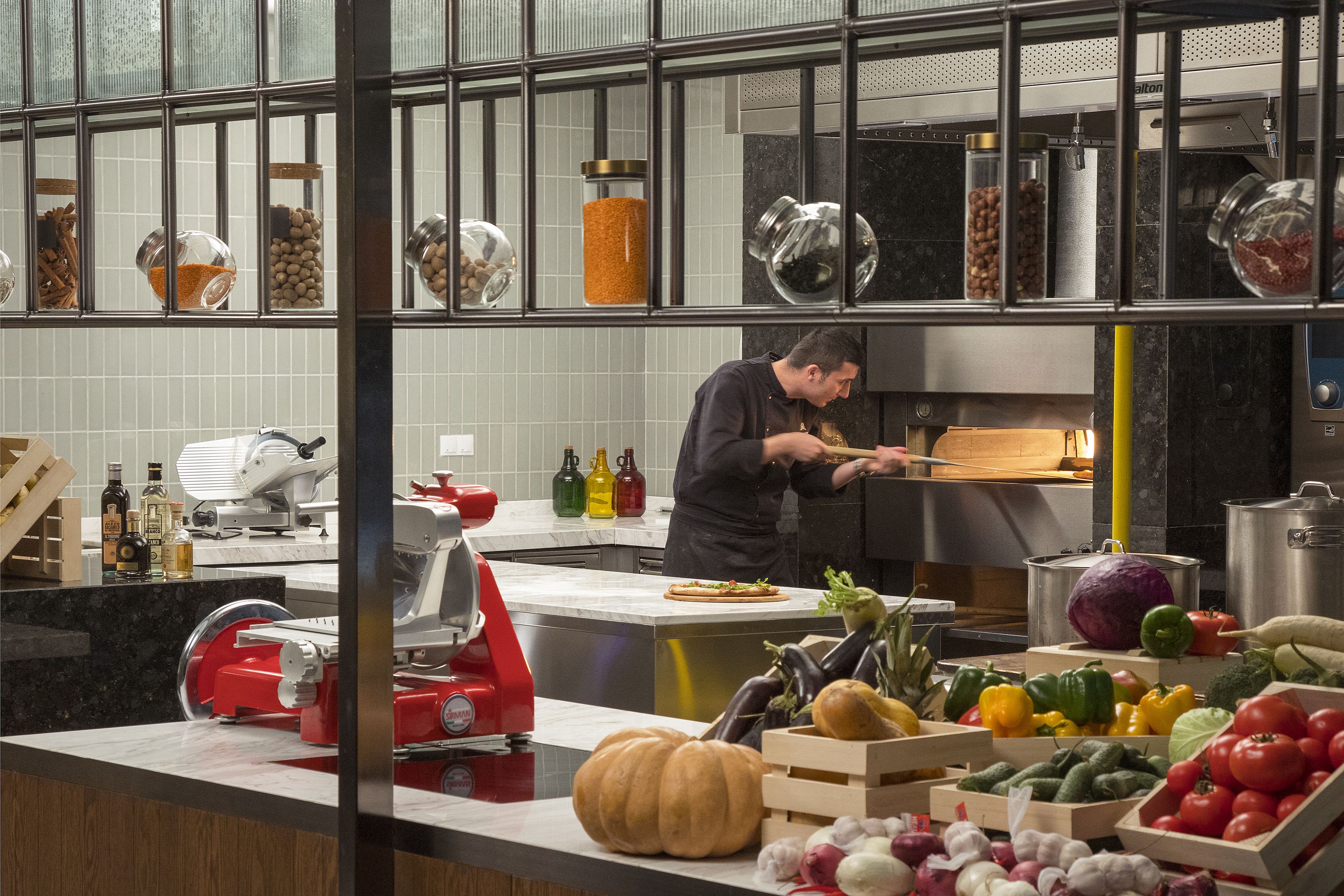 A kitchen with a chef preparing food, various kitchen appliances, and a display of fresh produce.