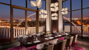 A dining area with a view of a cityscape at dusk, featuring a long table set for dining, large windows, and modern pendant lighting. - The St. Regis Astana (Astana)