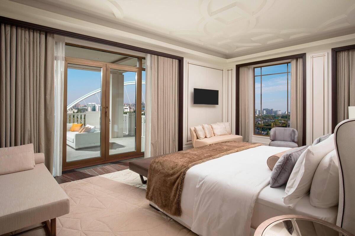Ein Hotelzimmer mit einem großen Bett, einem Sofa, einem Sessel, einem Fernseher und einem Balkon mit Blick auf die Stadt.