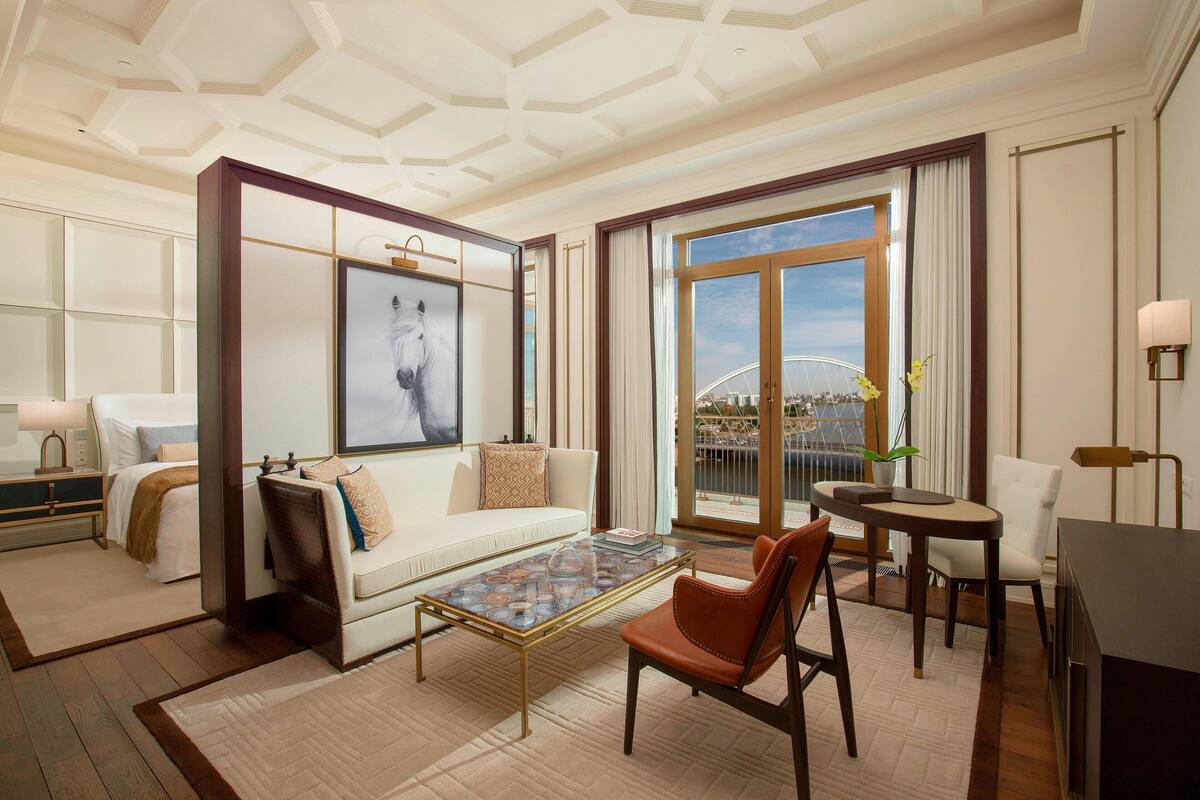 Ein geräumiges Hotelzimmer mit großem Fenster und Blick auf eine Brücke sowie die Stadt. Das Zimmer verfügt über eine Couch, einen Couchtisch und einen Essbereich mit einem Stuhl.