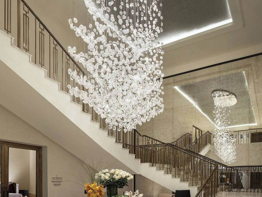 A grand staircase with a modern chandelier, marble flooring, and a seating area with a table and chairs.