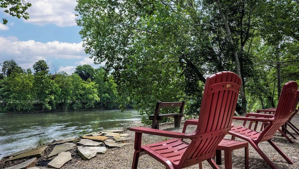 Outdoor seating with Adirondack chairs near a river, surrounded by trees.
